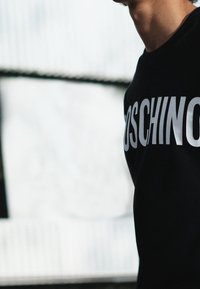 Černá mikina s bílým logem "Moschino" výrazně vytištěným na hrudi, klasického střihu a měkké textury látky.