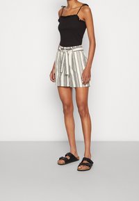 Haut noir ajusté avec bordure à volants, associé à un short crème avec des rayures noires à motif. Porte des sandales noires à enfiler.