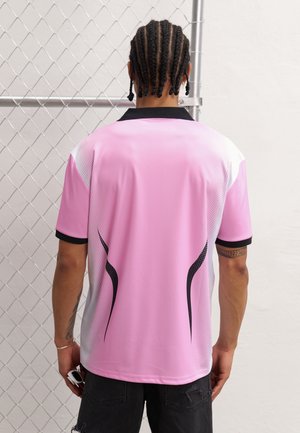 Person mit geflochtenem Haar steht mit dem Gesicht zu einem Maschendrahtzaun, trägt ein rosa-weißes Kurzarmshirt mit schwarzen Akzenten und schwarze Shorts.
