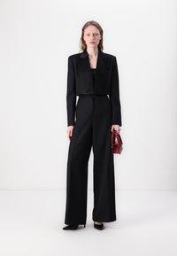 Musta tuxedo-takki satiinikaulukalla, lyhyt malli, yhdistettynä korkeavyötäröisiin mustiin housuihin, pienen burgundilaukun kanssa ja mustissa korkokengissä.