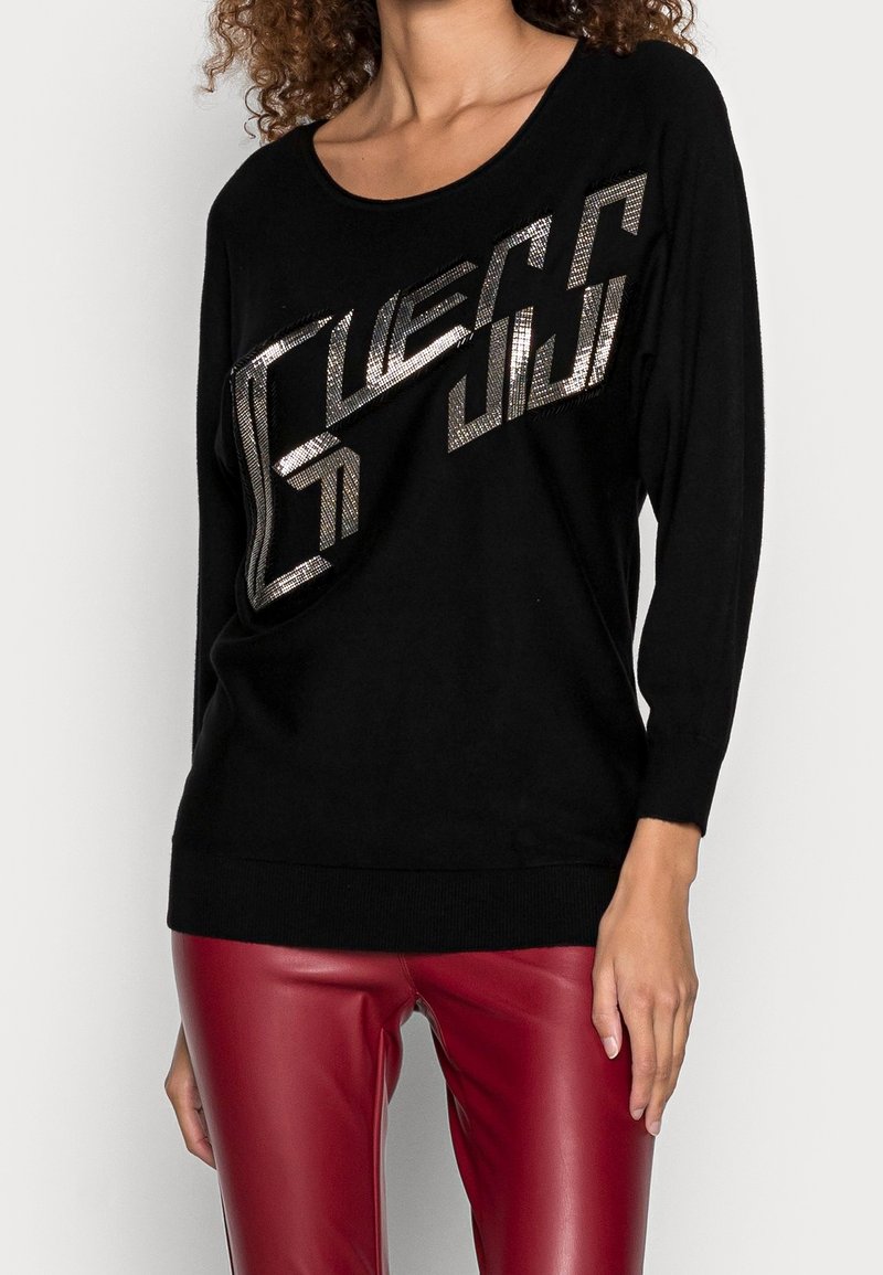 Femme portant un haut noir à manches longues avec un texte géométrique brillant et un pantalon en cuir rouge sur un fond clair uni.