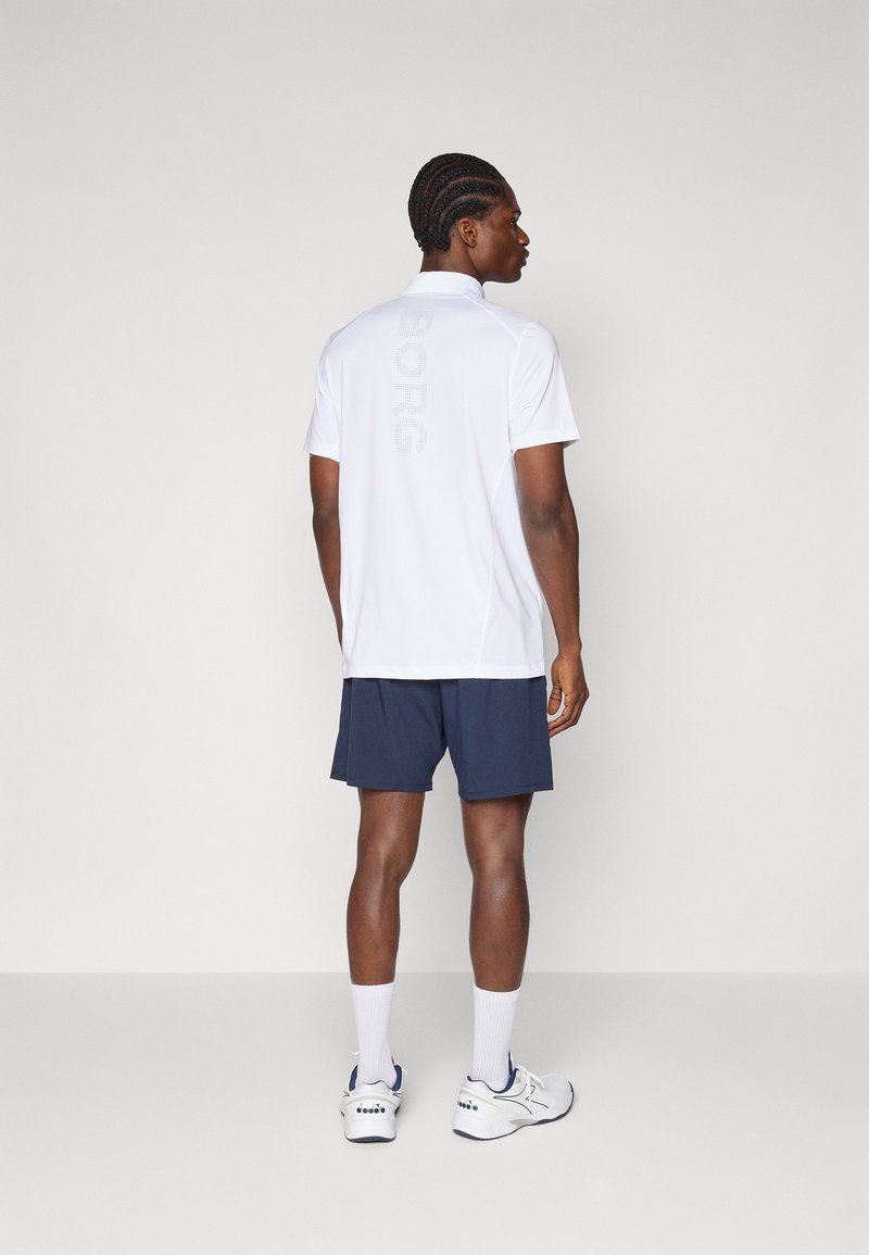 Camisa polo branca com discreta marca "BORG", combinada com calções azuis-marinho. Modelo usa ténis de desporto e meias brancas, visto de trás.