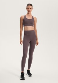 Atletski set u umirujućoj mauve boji, sastavljen od prilagođenog kratkog topa i visokih tajica. Glatka tekstura s minimalnim šavovima, uparen s crnim tenisicama.