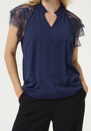 Donna che indossa una camicetta blu navy con maniche corte in pizzo e colletto arricciato, abbinata a pantaloni neri, mani nelle tasche.