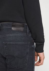 Jeans de ganga escuros com um acabamento texturizado, apresentando um patch de marca em pele na cintura e um bolso traseiro com costura curva.