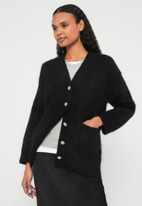 Μαύρη ζακέτα από χνουδωτό υλικό, με V-λαιμόκοψη, μακριά μανίκια, μεγάλα κουμπιά με διακοσμητικά στοιχεία και πλαϊνή τσέπη.