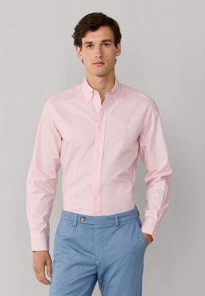 Mann trägt ein hellrosa Hemd mit Knöpfen, das in hellblaue Hosen gesteckt ist, und steht vor einem grauen Hintergrund.