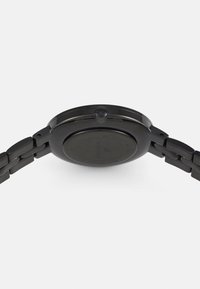 Orologio analogico rotondo di colore nero con bracciale in metallo, dotato di una lunetta liscia e di una corona di carica texturizzata. Sul retro presenta dettagli di branding minimi.