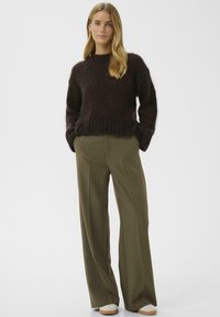 Tumši brūns, pūkains adīts džemperis ar ribotu apakšmalu, kas tiek kombinēts ar augsta vidukļa plašām olīvu zaļām biksēm. Ikdienišķa, teksturēta apavu pāris noslēdz izskatu.