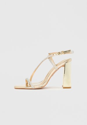 Goldener High-Heel-Sandalette mit glänzender Lackoptik, transparenten Strassriemen und klobigem Blockabsatz. Quadratische Zehenpartie.