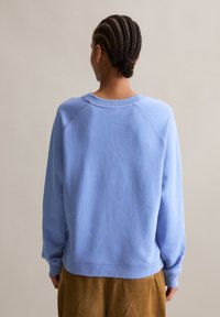Persona con un chignon intrecciato, che indossa un maglione azzurro chiaro e pantaloni marroni, vista di spalle su uno sfondo neutro.