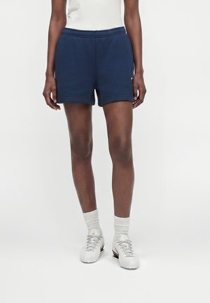 Person iført marineblå shorts, hvide crew-sokker, hvide snørebåndssneakers og en hvid ærmeløs top mod en ensfarvet hvid baggrund.