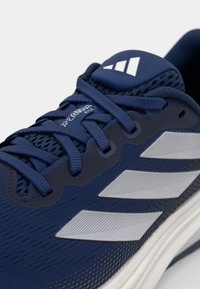 Zapatilla atlética azul con parte superior de tela, diseño texturizado, cordones en azul tonal, tres franjas blancas y un logo en la lengüeta acolchada.