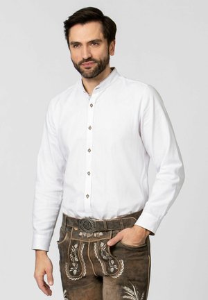 Weißes Langarmhemd mit Stehkragen, versehen mit dekorativen Knöpfen. Kombiniert mit braunen Lederhosen, geschmückt mit weißer Blumenstickerei.