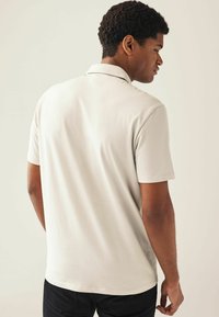 Camisa polo beige con una textura suave, de mangas cortas y un collar clásico, vista desde la parte posterior, combinada con pantalones negros.