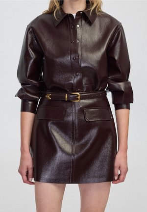 Femme portant une robe en cuir brun foncé brillant avec des manches longues, des boutons-pression, deux poches avant et une ceinture assortie avec une boucle dorée.