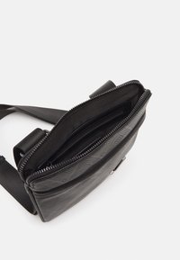 Svart syntetisk crossbody-väska med geometriskt mönster, dragkedjestängning och justerbar rem. Insidan har flera fack för organisation.