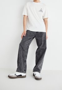 Camiseta blanca de algodón con un pequeño triángulo negro y diseño de ojo, combinada con unos jeans de mezclilla grises de corte suelto y zapatillas blancas con acentos negros.