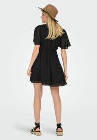Robe noire courte avec des manches courtes transparentes, taille cintrée et jupe fluide. Associée à un chapeau à large bord beige et des sandales noires.
