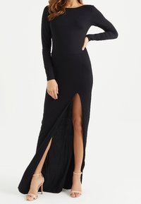 Kvinna klädd i en lång svart klänning med långa ärmar och en hög framklippning, tillsammans med nudefärgade högklackade sandaler.