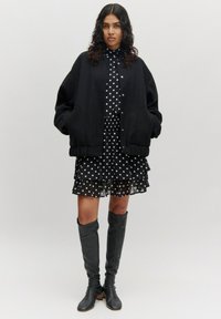 Zwarte bomberjack over een zwart polka dot jurk met ruches. Kniehoge zwarte leren laarzen. Steil haar los gedragen.