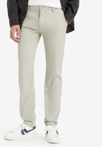 Pantaloni grigi chiari con una vestibilità aderente, realizzati in tessuto misto cotone. Presentano una chiusura con bottone e tasche laterali, abbinati a sneakers bianche.