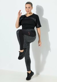 Kobieta w czarnym i w panterkę stroju sportowym podnosi jedno kolano i unosi jedną rękę, stojąc przy białej ścianie z cieniem.