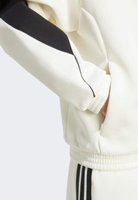 Veste de sportive couleur crème avec une bande noire sur la manche, tissu texturé, poignets côtelés et coupe ajustée à la taille.