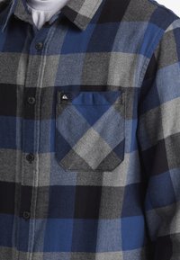 Blauw en grijs geruite flanellen overhemden met een knoopsluiting aan de voorkant, lange mouwen en een borstzak met een klein logo. Zachte textuur.