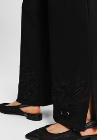 Zwarte wijde broek met bloemvormige uitsnijdingen aan de onderkant en zijsplitsingen. Gecombineerd met zwarte schoenen met puntige neuzen en een gestructureerd ontwerp.