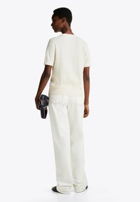 Maglione lavorato a maglia color crema a maniche corte con motivo a trecce, abbinato a pantaloni bianchi a gamba larga. Il modello tiene in mano una piccola borsa nera.