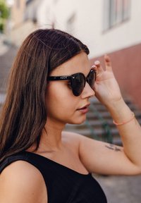 Černé sluneční brýle ve stylu kočičích očí mají lesklý povrch s nápisem značky "VUCH" na boku. Modelka má dlouhé vlasy a nosí černý top bez rukávů.