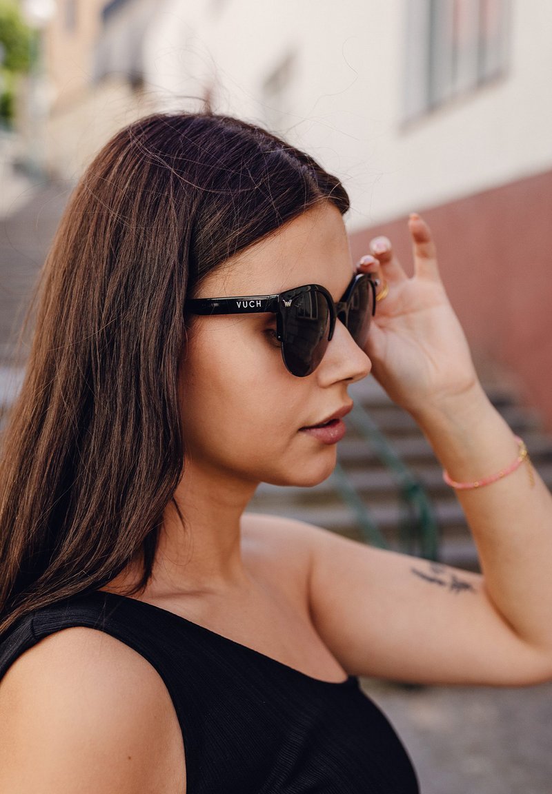 Černé sluneční brýle ve stylu kočičích očí mají lesklý povrch s nápisem značky "VUCH" na boku. Modelka má dlouhé vlasy a nosí černý top bez rukávů.