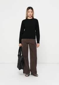 Svart långärmad tröja, bruna vida byxor i kordflort, och en svart tote-väska. Hightop sneakers med texturerad yta. Neutral bakgrund.
