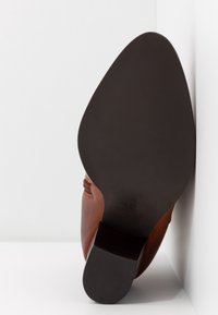 Semelle d'une chaussure en cuir marron avec une semelle noire lisse et un talon carré, appuyée verticalement contre un mur et une surface blancs.