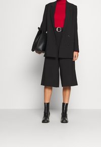 Femme en blazer noir, short ample longueur genou, bottes noires, col roulé rouge, tenant un grand sac à main noir sur un fond uni.
