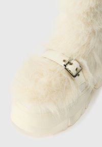 Plyšové bílé kotníkové boty s tlustou smetanovou platformou, s bílým koženým páskem a stříbrnou sponou. Měkký, strukturovaný vzhled.