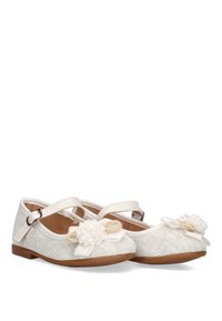 Zapatos de tela blanca con una superficie texturizada, que presentan una puntera redondeada, correa en el tobillo y detalles florales en la parte delantera. Suela plana marrón.