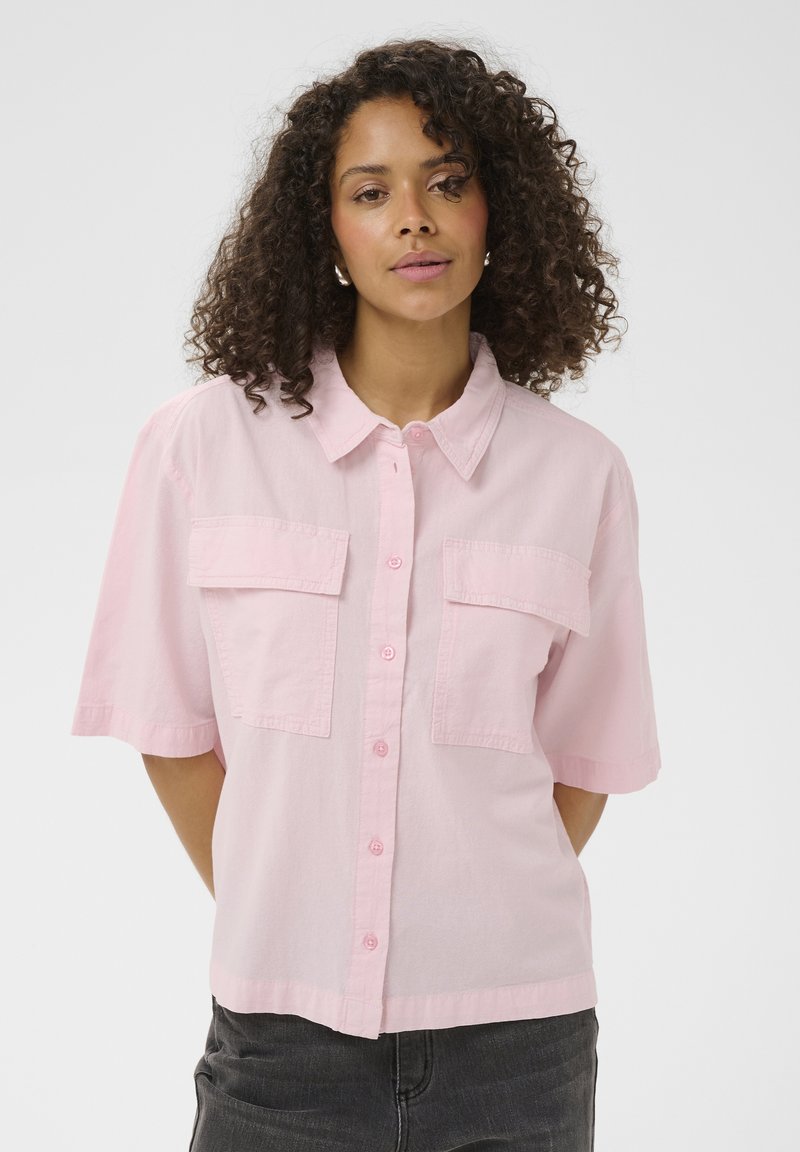 Femme aux cheveux bouclés portant une chemise rose clair à manches courtes avec deux poches avant, debout devant un fond blanc uni.
