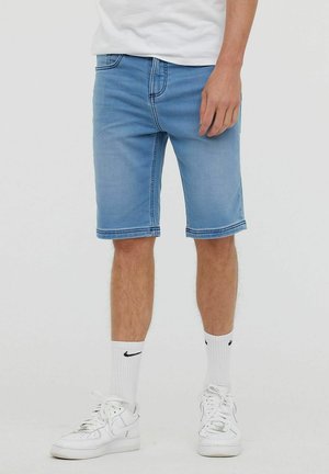 Shorts en denim bleu clair avec une coupe droite, des ourlets repliés et des coutures visibles. Portés avec des baskets blanches et des chaussettes.