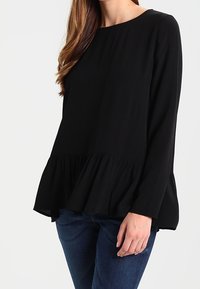 Blusa nera a maniche lunghe con scollatura tonda e orlo a peplo svasato. Tessuto morbido con vestibilità comoda, abbinata a jeans blu.