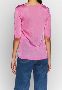 Top rosa a maniche corte con una texture liscia e leggermente trasparente. Presenta un collo rotondo e una silhouette aderente, indossato con jeans in denim blu.