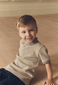 Polo de color beige con un patrón y un cuello marrón, con tres botones, combinado con pantalones azul oscuro. Niño sentado sobre un suelo de madera clara.