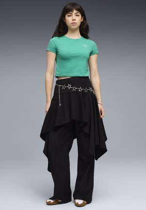 Frau trägt ein grünes bauchfreies Top, schwarzen asymmetrischen Rock über weiten Hosen, Stern-Ketten-Gürtel und weiße Sneakers mit brauner Sohle, steht vor grauem Hintergrund.