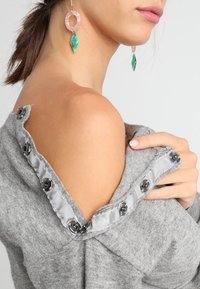 Šedý svetr s texturovaným povrchem a designem na jedno rameno. Obsahuje černé knoflíky podél výstřihu s pruhovaným lemem.