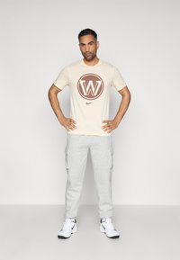 Béžové tričko s hnedým kruhovým logom a dizajnom "W", kombinované so svetlosivými cargo teplákmi a bielymi športovými topánkami.