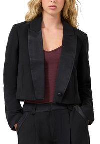 Sieviete, kas valkā melnu īsu žaketi ar satīna apmali, virs bordo krāsas V veida kakla topa un atbilstošām melnām biksēm ar satīna sānu svītrām.