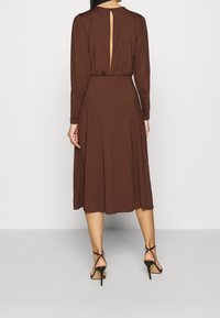 Robe longue à manches longues marron avec un détail en trou de serrure au dos, en tissu léger, avec un bas fluide, associée à des talons squelettes noirs.