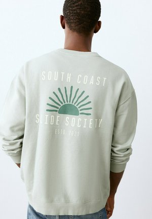 Person wearing light gray sweatshirt with green sun graphic and text "South Coast Slide Society Estd. 2021" on the back.