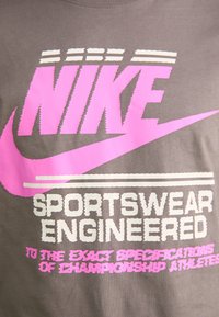 Pilkas marškinėliai, kuriuose dideli rožiniai ir balti užrašai: "NIKE" ir "SPORTSWEAR ENGINEERED." Dizainą papildo Nike swoosh logotipas.